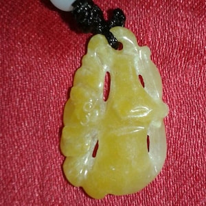 May include: A yellow jade pendant with a carved design, suspended from a black cord and white beaded necklace. The pendant has a smooth, polished surface and is set against a red background.