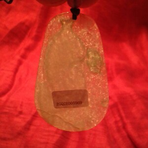 May include: A translucent, teardrop-shaped pendant with a black cord. The pendant has a sticker with the text "ZQQ2E005969". The background is a red fabric.