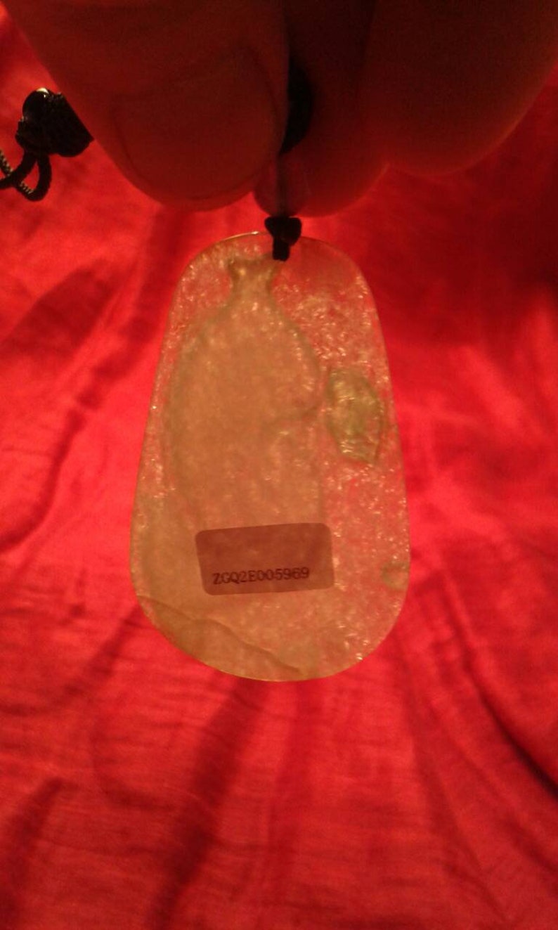 May include: A translucent, teardrop-shaped pendant with a black cord. The pendant has a sticker with the text "ZQQ2E005969". The background is a red fabric.