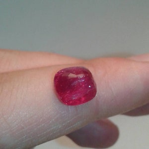 May include: A faceted, cushion-cut ruby gemstone. The gem is a vibrant red color and appears to be held by a person's finger. The ruby's facets reflect light, creating a sparkling effect. Ideal for jewelry making.