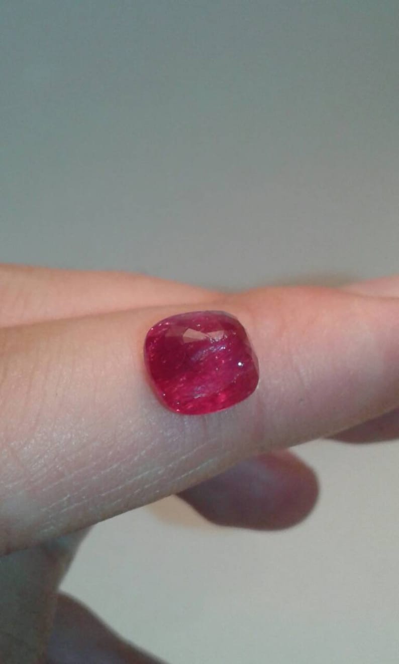 May include: A faceted, cushion-cut ruby gemstone. The gem is a vibrant red color and appears to be held by a person's finger. The ruby's facets reflect light, creating a sparkling effect. Ideal for jewelry making.