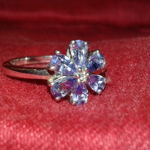 May include: A silver ring featuring a flower design with six light blue teardrop-shaped gemstones. The ring is set against a red fabric background, showcasing the ring's intricate details and color contrast.