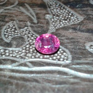 May include: A round, pink gemstone, possibly a sapphire or tourmaline, sits on a textured, dark surface. The gem's colour varies from light to deep pink, with internal facets reflecting light. The background has a decorative pattern.