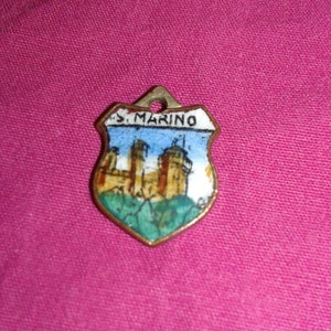 Pode incluir: Um pingente em forma de escudo com uma borda dourada e uma pequena argola na parte superior. O pingente apresenta uma cena pintada de um castelo com o texto "S. MARINO" no topo, contra um céu azul e uma paisagem verde.
