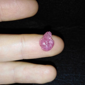 May include: A small, rough-cut, pink ruby gemstone, approximately 1.3 cm in length, rests on a fingertip. The ruby has a textured surface and a natural, organic shape, with a dark background.