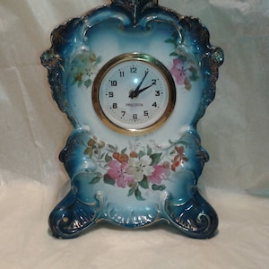 Puede incluir: Un reloj antiguo de porcelana con un diseño floral azul y blanco. La esfera del reloj es blanca con números negros y ribete dorado. La palabra "PRECISTA" está impresa en la esfera. El reloj tiene una forma decorativa y ornamentada.