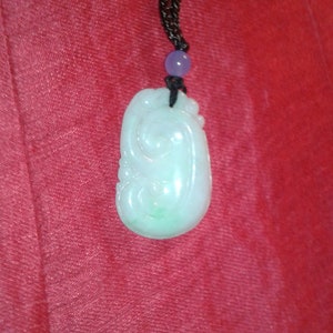 May include: A jade pendant necklace featuring a carved, pale green and white stone. The pendant hangs from a black cord with a purple bead. The backdrop is a plain red.