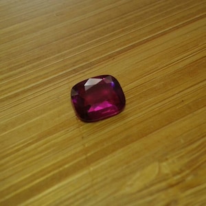 May include: A faceted, cushion-cut gemstone with a deep magenta hue. The gem is set against a warm, wooden surface, highlighting its brilliance and color. The gemstone's shape and color suggest it could be a ruby or similar precious stone.