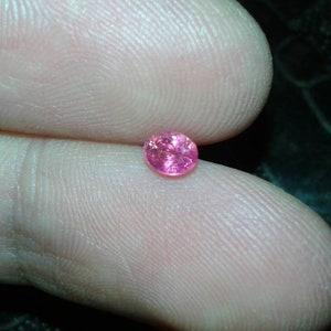 May include: A small, oval-shaped, pink gemstone. The gem is held between two fingers, showcasing its vibrant color and facets. The background is dark, highlighting the gem's brilliance. This gemstone is likely for jewelry making.