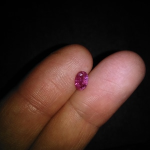 Peut inclure: Une petite pierre précieuse rose de forme ovale. La gemme est tenue entre deux doigts sur un fond sombre. Les facettes de la pierre précieuse reflètent la lumière, lui donnant un aspect scintillant. La gemme est probablement un rubis ou un saphir rose.