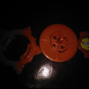 Pode incluir: Uma variedade de pe&ccedil;as de brinquedos de pl&aacute;stico laranja, incluindo uma pe&ccedil;a circular com um centro em forma de cruz, um anel pontiagudo e uma pequena pe&ccedil;a vermelha com um adesivo amarelo. Os itens est&atilde;o em uma superf&iacute;cie escura.