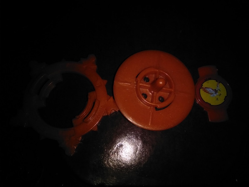Pode incluir: Uma variedade de pe&ccedil;as de brinquedos de pl&aacute;stico laranja, incluindo uma pe&ccedil;a circular com um centro em forma de cruz, um anel pontiagudo e uma pequena pe&ccedil;a vermelha com um adesivo amarelo. Os itens est&atilde;o em uma superf&iacute;cie escura.