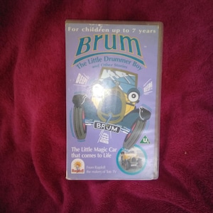 Puede incluir: Una cinta VHS para niños con el personaje de dibujos animados Brum. La portada es morada con el título "Brum" en letras azules grandes. La cinta incluye "The Little Drummer Boy and Other Stories". También se ve el texto "The Little Magic Car that comes to Life".