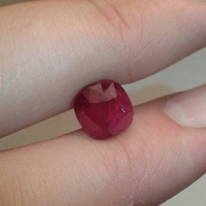 May include: A faceted, deep red gemstone, likely a ruby, held between fingers. The gem has a rounded shape with multiple facets catching the light, showcasing its rich color and clarity. The background is a neutral, blurred tone.