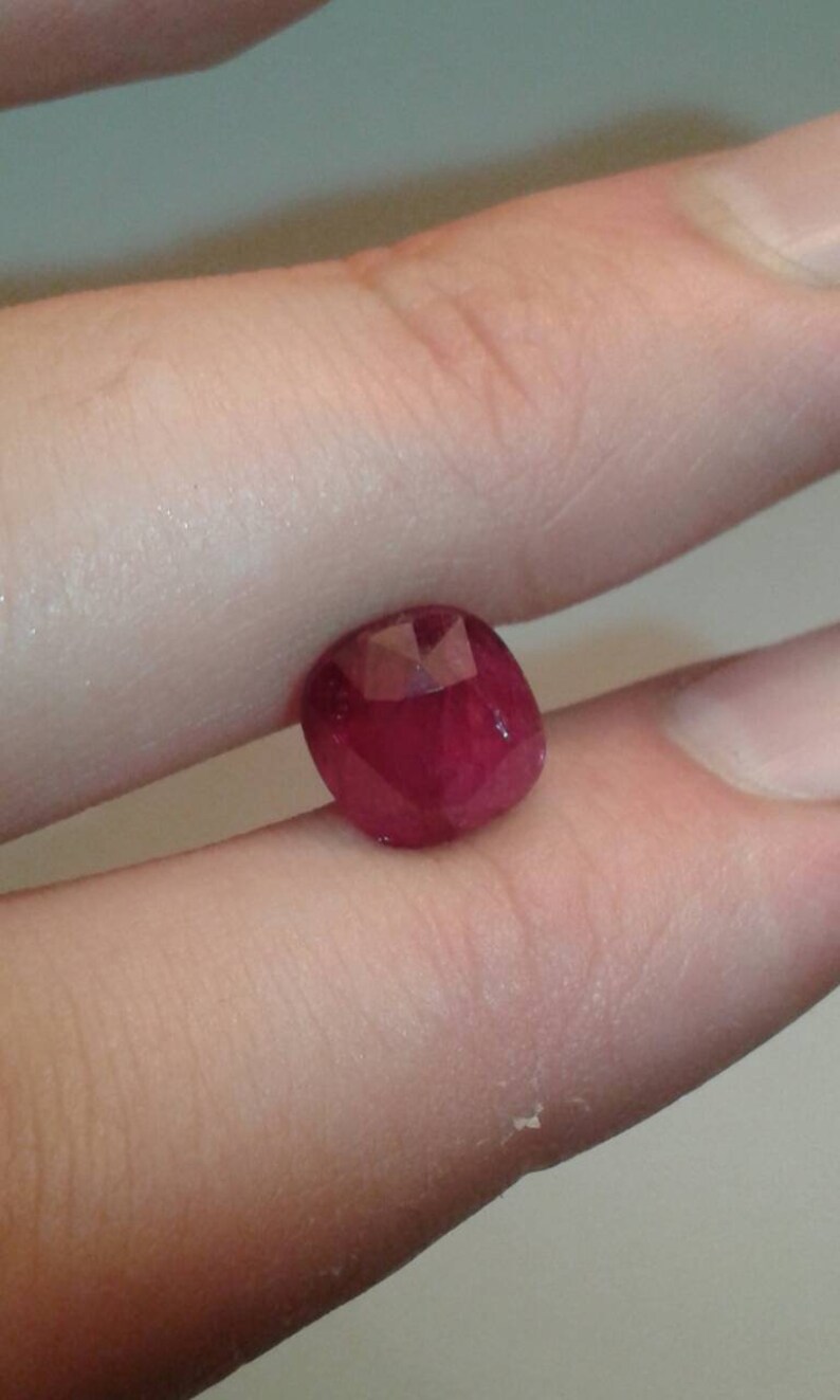 May include: A faceted, deep red gemstone, likely a ruby, held between fingers. The gem has a rounded shape with multiple facets catching the light, showcasing its rich color and clarity. The background is a neutral, blurred tone.
