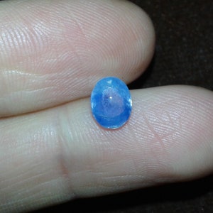 May include: A small, oval-shaped, light blue gemstone. The gem is held between two fingers, showcasing its facets and clarity. The stone's color is a vibrant, clear blue, reflecting light. The background is a dark brown.