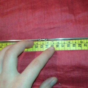 May include: A silver metal object, approximately 10 inches long, is placed next to a yellow measuring tape. The object has a twisted design in the middle and tapers to a point at one end. The background is a solid red fabric.