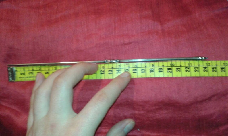 May include: A silver metal object, approximately 10 inches long, is placed next to a yellow measuring tape. The object has a twisted design in the middle and tapers to a point at one end. The background is a solid red fabric.