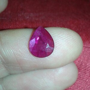 May include: A faceted, teardrop-shaped ruby gemstone. The gem is a deep, vibrant pinkish-red color, with multiple facets catching the light. The ruby is held between fingers, against a red background.