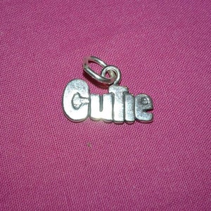 Op de afbeelding: Een zilverkleurige bedel met het woord "Cutie" in een vet, afgerond lettertype. De bedel heeft een kleine lus aan de bovenkant voor bevestiging en ligt op een roze achtergrond.