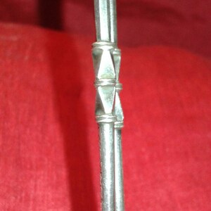 May include: Close-up of a silver metal object with a decorative geometric design. The object has a series of parallel lines and a diamond-shaped pattern in the center. The background is a solid red color.