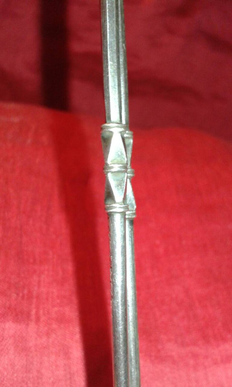 May include: Close-up of a silver metal object with a decorative geometric design. The object has a series of parallel lines and a diamond-shaped pattern in the center. The background is a solid red color.