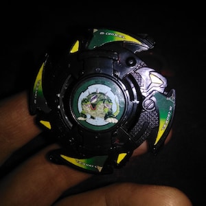 May include: A black and green toy with a circular design. The toy has green and yellow accents, with the text "M-DRAGEL" and a circular image of a green creature. The center of the toy has the text "MT 02".