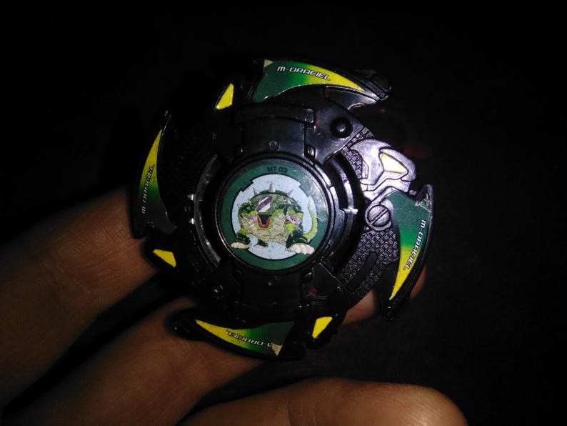 May include: A black and green toy with a circular design. The toy has green and yellow accents, with the text "M-DRAGEL" and a circular image of a green creature. The center of the toy has the text "MT 02".