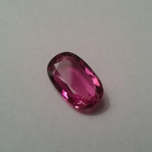 May include: A faceted, oval-shaped gemstone with a deep pink hue. The gem has a polished surface that reflects light, creating a sparkling effect. The stone is set against a plain white background.