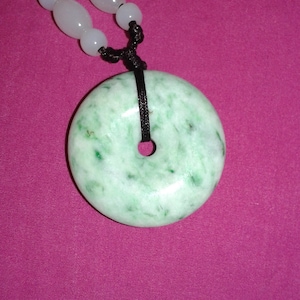 May include: A jade pendant necklace. The circular, light green stone has darker green speckles. It hangs from a black cord with white beads. The background is a magenta colour.