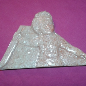 Puede incluir: Escultura triangular de color marrón claro de una persona con pelo largo y una túnica. La figura sonríe y sostiene un objeto. La escultura está hecha de un material similar a la piedra y está sobre un fondo púrpura.