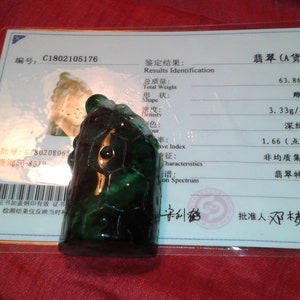 May include: A dark green jade turtle carving, presented on a red surface. The carving is accompanied by a certificate with Chinese text, detailing the jade's weight and density in grams.