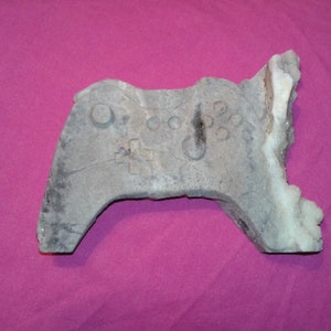 May include: A gray, stone-like sculpture of a video game controller with button details. The controller is set against a magenta background, with a crystalline formation on the right side.
