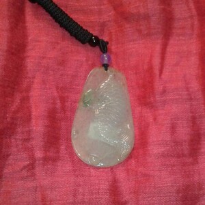 May include: A pale green jade pendant with a carved bird design, suspended from a black cord necklace. The pendant has a teardrop shape and is set against a red background.