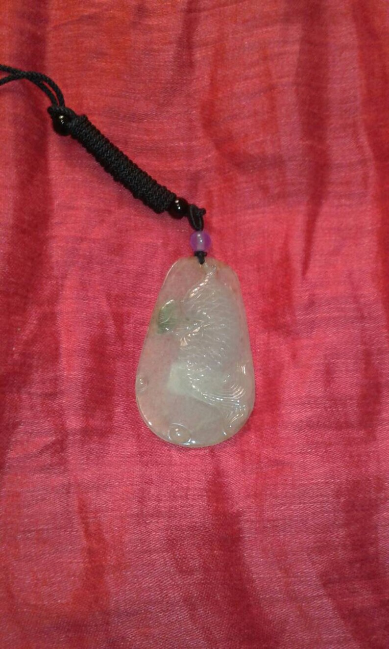 May include: A pale green jade pendant with a carved bird design, suspended from a black cord necklace. The pendant has a teardrop shape and is set against a red background.