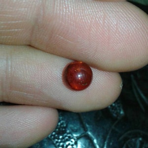 May include: A small, round, polished gemstone with a deep reddish-orange hue. The gem is held between two fingers, showcasing its smooth, reflective surface. The background is blurred, focusing attention on the gem's colour and form.