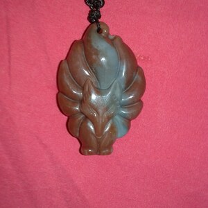 May include: A brown and blue fox pendant with a nine-tailed design, suspended from a black cord with a decorative knot. The pendant is carved with intricate details, showcasing the fox's features. The background is a solid red color.
