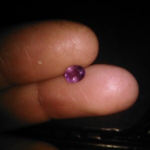 May include: A small, oval-shaped, purplish-pink gemstone. The gem is held between two fingers against a dark background, highlighting its color and shape. The stone appears to be polished and reflects light.
