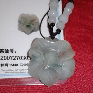 May include: A jade pendant necklace featuring a large, flower-shaped pendant with a smaller matching piece. The pendant is light green and white, with a black cord and white beads. The background is a red fabric.