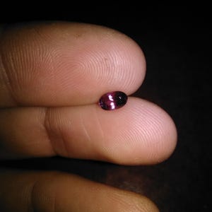 May include: A small, oval-shaped gemstone with a deep magenta hue, held between two fingers. The gem reflects light, showcasing its polished surface and internal color variations. The background is dark.