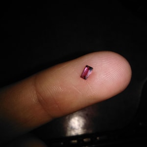 Puede incluir: Una pequeña gema rectangular y facetada con un tono rosa intenso, apoyada en la punta de un dedo. La gema tiene un interior oscuro, casi negro. El fondo es un espacio oscuro y borroso.