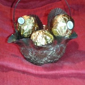 Puede incluir: Una cesta plateada con asa, llena de tres bombones envueltos en oro. La cesta tiene un diseño floral ornamentado y se encuentra sobre una superficie de tela roja. Los bombones tienen forma redonda y una pequeña etiqueta blanca.