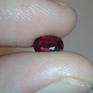 Peut inclure: Une pierre précieuse rouge foncé, de forme ovale et facettée, est tenue entre deux doigts. La gemme a une surface polie et reflète la lumière, mettant en valeur sa structure interne. La couleur est un rouge riche et saturé.