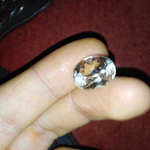 May include: An oval-shaped, clear gemstone with internal light reflections. The gem is held between fingers against a dark red background. The stone appears to be faceted, catching and reflecting light.