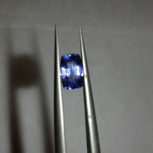 Könnte beinhalten: Ein kissenförmiger, tiefblauer Edelstein, der von einer silbernen Pinzette gehalten wird. Der Edelstein hat eine facettierte Oberfläche, die das Licht reflektiert und einen funkelnden Effekt erzeugt. Die Pinzette befindet sich vor einem neutralen Hintergrund.