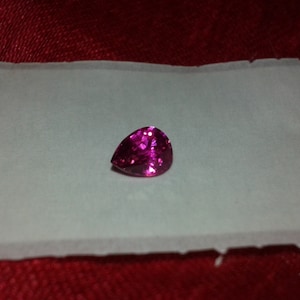 May include: A pear-shaped, faceted, magenta gemstone rests on a white surface against a red background. The gemstone has a brilliant, sparkling appearance, reflecting light. The setting is simple, highlighting the gem's color and cut.