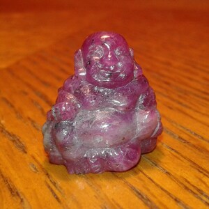 May include: A small, carved figurine of a smiling Buddha. The statue is a deep pink color with lighter pink and white accents. The Buddha is sitting in a relaxed pose, with a rounded belly and serene expression.