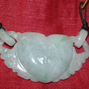 May include: A pale green jade pendant with a heart-shaped design and carved Chinese characters. The pendant is suspended on a gold and green cord, with a red background.