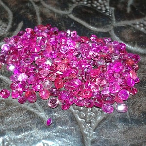 May include: A pile of faceted, pink gemstones of varying sizes. The gems are scattered on a dark, textured surface. The gemstones have a brilliant, reflective quality, with shades ranging from light to deep pink.