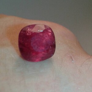 May include: A cushion-cut, deep red ruby gemstone. The gem is faceted, reflecting light, and appears to be set against a skin-toned background. The ruby's rich color and shape are the focal point.